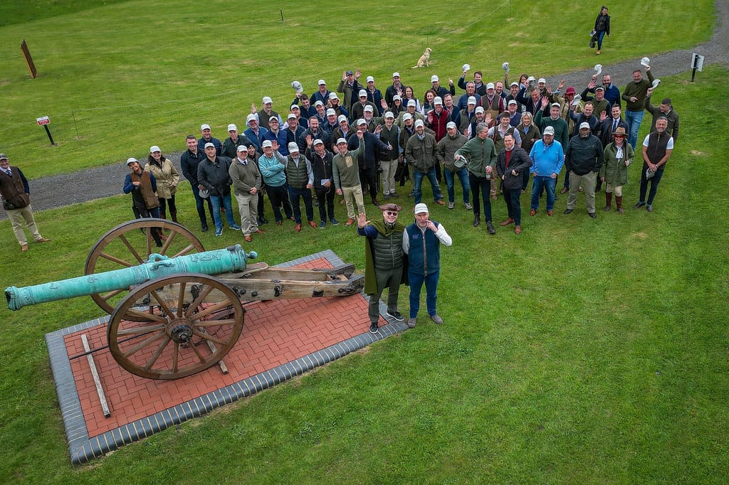 An aerial shot of a large group of people waving at the camera next to an ornamental cannon.