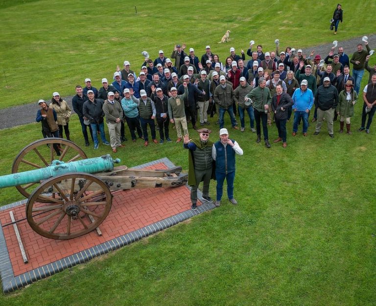An aerial shot of a large group of people waving at the camera next to an ornamental cannon.